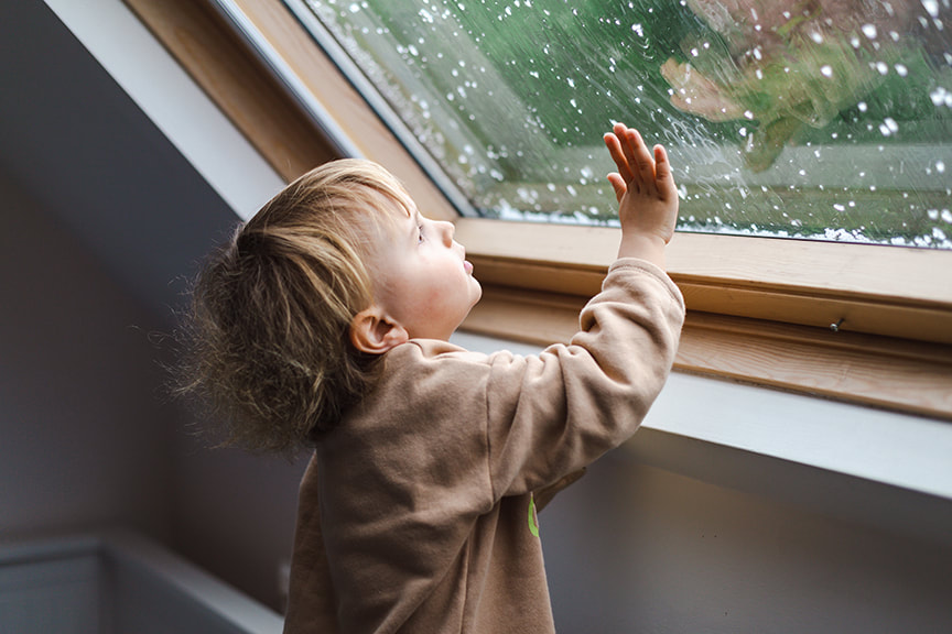 Little preschool boy staying home in bad weather and looking with interest through window on raindrops and hail, idoors.